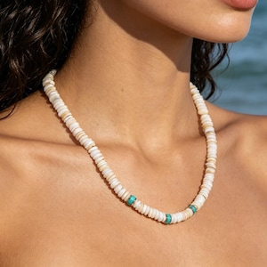 Könnte beinhalten: Eine Perlenkette bestehend aus einer Reihe kleiner, flacher, cremeweißer, kreisförmiger Muschelscheiben, die aneinandergereiht sind. Die Kette ist mit zwei kleinen, leuchtend türkisfarbenen zylindrischen Perlen akzentuiert, die symmetrisch angeordnet sind.
