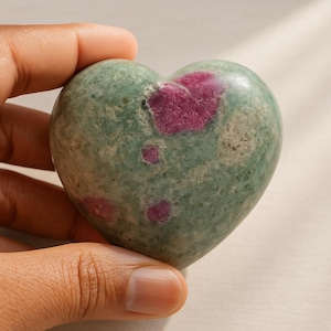 以下が含まれることがあります： 緑色のベースカラーとルビーレッドのパッチが特徴のハート型の石。滑らかで磨かれた表面が際立つように、石は手で持たれています。家の装飾やユニークなギフトに最適な装飾品です。