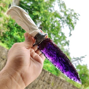 Könnte beinhalten: Ein dekorativer Dolch mit einer durchscheinenden, tiefvioletten, scharfkantigen Klinge mit strukturierter Oberfläche. Der Griff ist aus einem hellen, cremeweißen Material in organischer Form geschnitzt und durch ein silberfarbenes Metallparierstück mit aufwendigen, geprägten Schnörkelmustern mit der Klinge verbunden.