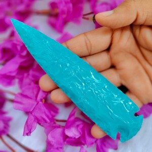Könnte beinhalten: Ein türkisfarbener Pfeilspitze mit einer scharfen Spitze und geschnitzten Details. Die Pfeilspitze wird in einer Hand gehalten, mit einem Hintergrund aus rosa Blumen.