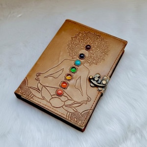 Può includere: Un diario in pelle marrone con un design in rilievo di una figura meditante e simboli dei chakra. Il diario presenta una chiusura in metallo ed è adornato con pietre preziose colorate che rappresentano i chakra.