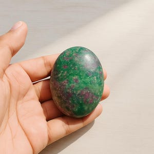 May include: A smooth, oval gemstone in shades of green and burgundy. The stone is held in a hand, highlighting its polished surface. The natural stone has a mottled pattern and a glossy finish. The stone is approximately 6 cm in length.
