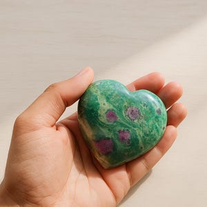 Może przedstawiać: Kamień w kształcie serca z zieloną podstawą i czerwonymi wtrąceniami. Kamień trzymany jest w dłoni, ukazując jego wypolerowaną powierzchnię i unikalny wzór kolorystyczny. Kamień ma około 7.6 cm szerokości.