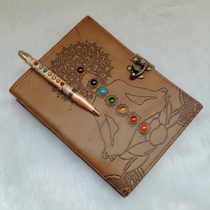 Può includere: Un diario in pelle marrone con un design mandala dettagliato e una chiusura. Una penna con pietre preziose colorate è appoggiata sul diario. Il diario presenta una figura incisa con simboli chakra.