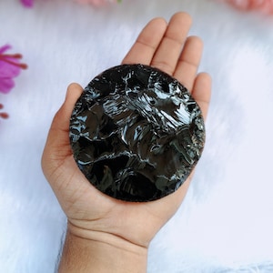Puede incluir: Una piedra de obsidiana negra pulida y redonda. La piedra tiene una superficie brillante y reflectante y un borde ligeramente irregular. El color oscuro y la textura suave sugieren un artículo natural y artesanal, posiblemente para fines decorativos o espirituales.