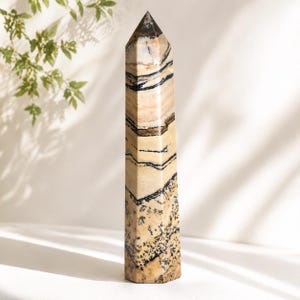 Pode incluir: Obelisco de pedra polida com topo pontiagudo. A pedra apresenta um padrão de faixas e manchas bege, creme e pretas. O obelisco é um objeto decorativo, provavelmente para decoração de interiores ou práticas espirituais.