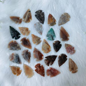 Puede incluir: Una colección de pequeñas puntas de flecha hechas a mano en varios colores, incluyendo marrón, beige, negro y verde. Las puntas de flecha son de piedra y están dispuestas sobre una superficie blanca y esponjosa. Estas puntas de flecha decorativas son ideales para coleccionistas.