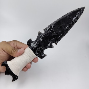 May include: A black obsidian dagger featuring a sharp, serrated blade with a glossy, glass-like finish. The handle is wrapped in a light beige, textured cord, and the base of the handle terminates in a pointed, black obsidian decorative element.