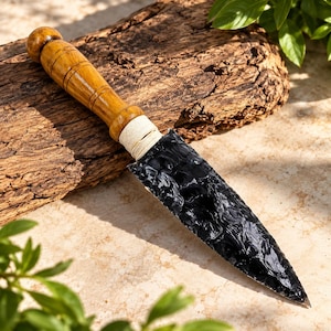 Peut inclure: Un poignard doté d'une lame tranchante en obsidienne noire à la texture brillante et écaillée. Le manche est fabriqué en bois brun clair poli avec des rainures horizontales, relié à la lame par un lien en corde blanc cassé serré.