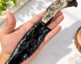 以下が含まれることがあります： 光沢のある黒い黒曜石の刃を持つナイフ。刃先は質感のある欠けた形状で、ハンドルは自然な茶色とクリーム色の鹿の角で作られており、表面は粗く有機的な質感で、根元は暗い色合いをしています。