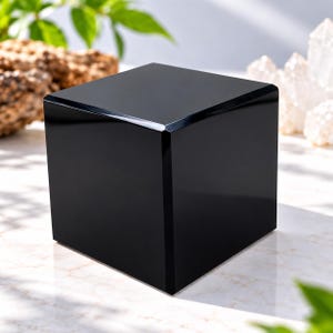 Könnte beinhalten: Ein massiver schwarzer Würfel mit einer glatten, reflektierenden und polierten Oberfläche. Das Objekt weist scharfe, klare Kanten und ein gleichmäßiges dunkles Finish auf und präsentiert eine geometrische Form mit glänzendem Aussehen.