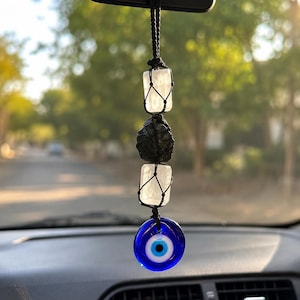 May include: A hanging car ornament featuring two rectangular white selenite crystals and one central rough black tourmaline stone, all encased in black macrame netting, finished with a circular blue glass evil eye bead at the bottom.