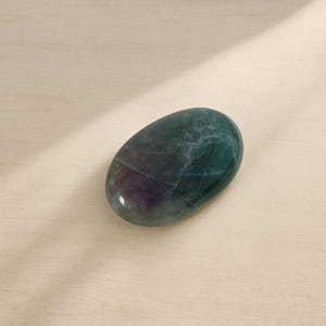 May include: A polished, oval-shaped stone with a smooth surface. The stone displays a mix of colours, including deep green, purple, and blue hues. The stone's surface has natural veining and colour variations, enhancing its unique appearance.