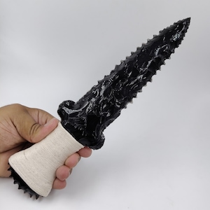 May include: A black obsidian dagger featuring a sharp, serrated blade with a glossy, glass-like finish. The handle is wrapped in a light beige, textured cord, providing a firm grip. The blade exhibits distinct, hand-knapped edges and a pointed tip.