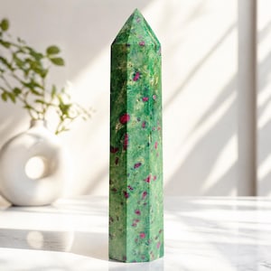 Puede incluir: Un obelisco de cristal verde con inclusiones rojas. La gema pulida tiene una forma rectangular facetada con una parte superior puntiaguda. El obelisco está sobre una superficie blanca. Un jarrón blanco y vegetación en el fondo.