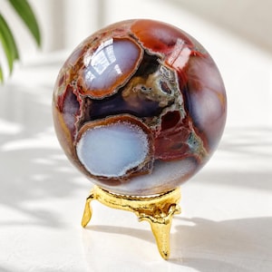 Puede incluir: Una esfera de piedra preciosa pulida, con una mezcla de colores como rojo, marrón y azul claro, se exhibe sobre un soporte de metal dorado. La esfera tiene una superficie lisa y reflectante con un patrón único. El soporte tiene un diseño ornamental. Diámetro aproximado: 10 cm.