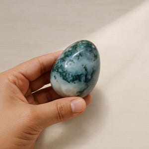 Puede incluir: Una piedra pulida en forma de huevo con una mezcla de patrones verdes y blancos. La piedra tiene una superficie lisa y brillante y se sostiene en una mano. Los colores y patrones crean una estética natural y orgánica. El objeto es decorativo.