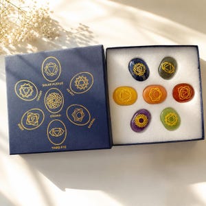 Op de afbeelding: Een donkerblauwe vierkante doos met goudkleurige chakra-symbolen en labels zoals Root, Throat, Solar Plexus, Heart, Crown, Sacral en Third Eye. Binnenin liggen zeven gladde, ovale stenen in diverse kleuren met bijpassende gouden gravures.