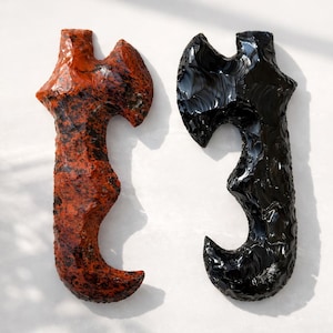 May include: Two decorative axe-shaped objects. One is a mottled red-orange color with black accents, the other is a glossy black. Both have a curved handle and a flared blade, resembling ancient tools.