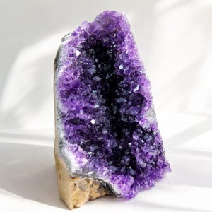 Könnte beinhalten: Eine große Amethyst-Geode mit tiefvioletten, kristallinen Strukturen, die in einer natürlichen, unebenen Gesteinsformation angeordnet sind. Das Innere zeigt einen leuchtenden, dunkelvioletten Farbton mit vielen Facetten.