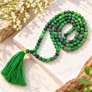 May include: A mala necklace featuring round green and black speckled gemstone beads with occasional small magenta inclusions. The necklace is finished with a vibrant green thread tassel secured by a small gold-coloured metal band at the top.