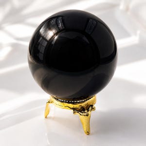 Puede incluir: Una gran esfera negra pulida, posiblemente obsidiana, descansa sobre un soporte dorado ornamental. La esfera refleja la luz, creando reflejos. El soporte tiene tres patas decorativas. El conjunto sugiere un objeto decorativo o espiritual. Diámetro aproximado de 10 cm.