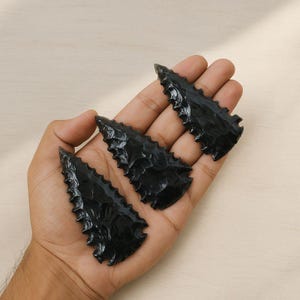 May include: Three black obsidian arrowheads with serrated edges are displayed in a hand. The arrowheads have a triangular shape and a glossy finish. The background is a light beige color.