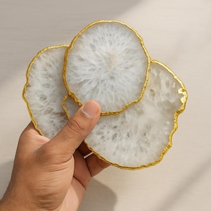 May include: Three white agate coasters with gold gilded edges. The coasters have a natural, organic shape and a translucent appearance. The gold trim adds a touch of elegance to the coasters, which are held in a hand.