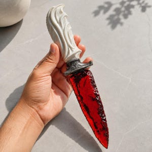 Pode incluir: Uma adaga decorativa com uma lâmina vermelha translúcida e serrilhada com superfície texturizada. O cabo é branco com padrões esculpidos em espiral e é separado da lâmina por uma guarda de metal escuro ornamentada com detalhes em filigrana.