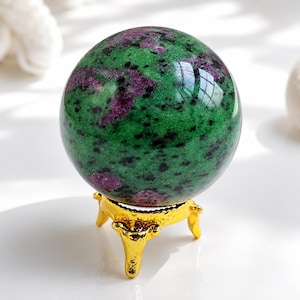 May include: A polished gemstone sphere, primarily green with deep red and black inclusions, sits on a small, ornate gold-colored stand. The sphere reflects light, showcasing its smooth surface and intricate patterns. A decorative item.