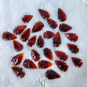 Puede incluir: Colección de piedras translúcidas en forma de punta de flecha, de color marrón rojizo. Las piedras están dispuestas sobre una superficie blanca y esponjosa. Las puntas de flecha tienen un acabado brillante y tamaños variables.