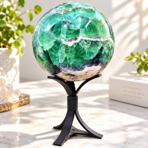 Pode incluir: Uma grande esfera de cristal de fluorita polida com tons vibrantes de verde esmeralda, azul-petróleo e roxo profundo, apoiada em um suporte de tripé de metal preto. A pedra exibe fraturas internas naturais e inclusões minerais brancas por toda.