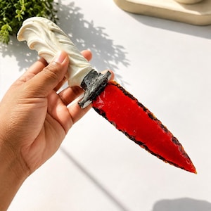 May include: A decorative dagger featuring a translucent, vibrant red blade with a jagged, textured edge. The handle is off-white with a sculpted, swirling pattern and is separated from the blade by a dark, weathered, metallic-looking bolster.