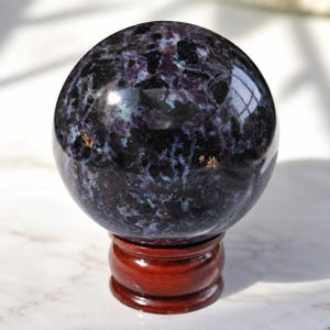 Puede incluir: Una esfera de piedra preciosa pulida, oscura y moteada, con tonos negros, morados y toques de azul. La esfera descansa sobre un pequeño soporte redondo de madera marrón rojizo, creando contraste.