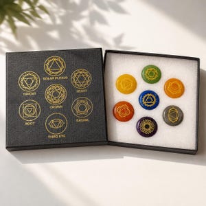 May include: A black square box containing seven circular stones in various colours, each engraved with a gold chakra symbol. The box lid features gold text labels for the Root, Sacral, Solar Plexus, Heart, Throat, Third Eye, and Crown chakras.
