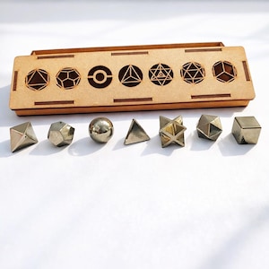以下が含まれることがあります： 蓋に幾何学模様がカットされた木製の箱と、その下に金属製の幾何学模様のセット。形状には、ピラミッド、球体、立方体、その他の多面体が含まれます。金属の形状は銀色と金色です。