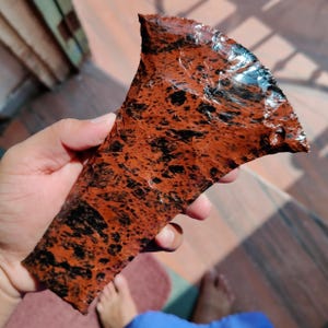 May include: A piece of red-brown obsidian with black speckles. The obsidian has a glossy, reflective surface and an irregular shape. The stone is held in a hand, showcasing its natural texture and color variations.