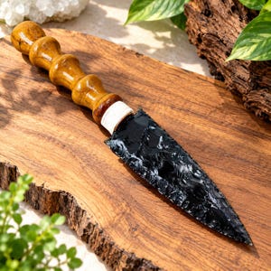 Peut inclure: Un poignard doté d'une lame en obsidienne noire tranchante à la texture brillante et écaillée. Le manche est fabriqué en bois brun clair poli avec une série de segments arrondis et bombés. Un cordon blanc est enroulé autour de la base.