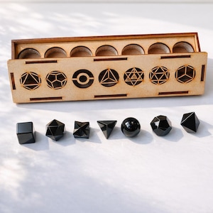 Könnte beinhalten: Eine Holzkiste mit sieben geometrischen Formen, die in die Vorderseite geschnitten sind, und sieben schwarzen Obsidiankristallformen darunter. Die Kiste hat kreisförmige Aussparungen für jeden Kristall. Die Kristalle umfassen einen Würfel, einen Stern, eine Kugel und andere Polyeder.