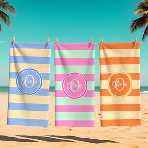 May include: Three personalized beach towels hanging on a clothesline. Each towel has horizontal stripes in different colors: blue and yellow, pink and mint, and orange and yellow. Each towel features a circular design with the name "Olivia".
