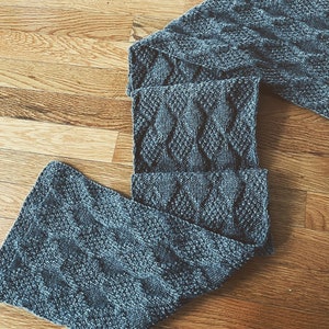 May include: A grey knitted scarf with a cable knit pattern. The scarf is folded in half and laid on a wooden surface.