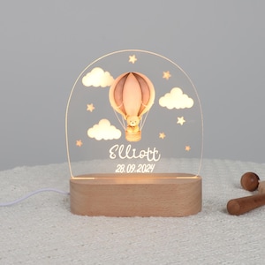 Pode incluir: Uma luz noturna personalizada com um design de balão de ar quente, um ursinho de pelúcia, nuvens e estrelas. O nome "Elliott" e a data "28.09.2024" são exibidos no painel de acrílico, que fica em uma base de madeira.