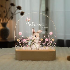 May include: A decorative night light featuring a bunny holding flowers, with the name "Sabrina" and the date "09.20.2021" printed on the clear acrylic. The light sits on a wooden base and is surrounded by floral and butterfly designs.