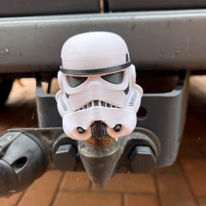 May include: A white Stormtrooper helmet-shaped trailer hitch cover. The cover has black detailing around the eyes and mouth, and is mounted on a black trailer hitch. The background shows a vehicle's bumper.