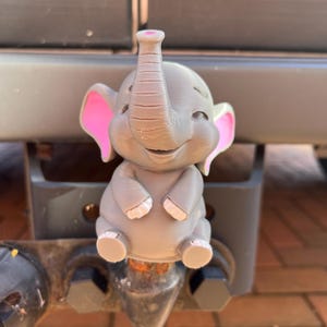 May include: A grey, 3D-printed elephant ornament with pink inner ears and toenails. The elephant has a smiling face and its trunk is raised. The ornament is attached to a trailer hitch.
