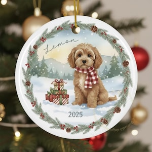Puede incluir: Adorno navideño redondo de cerámica con un perro marrón que lleva una bufanda a cuadros rojos y blancos, en un paisaje nevado con abetos. El adorno tiene la palabra "Lemon" y el año "2025". También hay una pila de regalos envueltos.