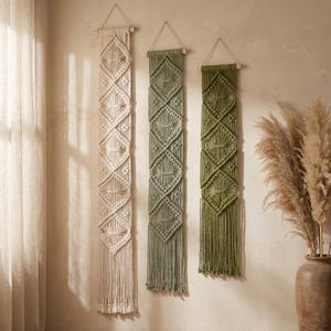 May include: Three macrame wall hangings in cream, sage green, and olive green. Each piece features a diamond pattern and fringe. They are suspended from wooden dowels with a hanging cord. The wall hangings are a bohemian style.