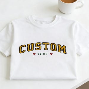 May include: White t-shirt with the word "CUSTOM" in gold block letters with a black outline. Below the word "CUSTOM" is the word "TEXT" in green, with a red heart on each side. A white mug with coffee sits in the background.