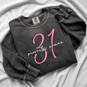 May include: Dark gray sweatshirt with the text "31 proverbs woman" in pink and white. The sweatshirt is folded on a white surface. The sweatshirt has a crew neck and long sleeves.
