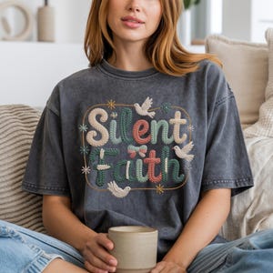 May include: A dark gray t-shirt with the embroidered text "Silent Faith" in colorful letters. The design includes floral and bird accents. The shirt has a vintage, washed appearance.